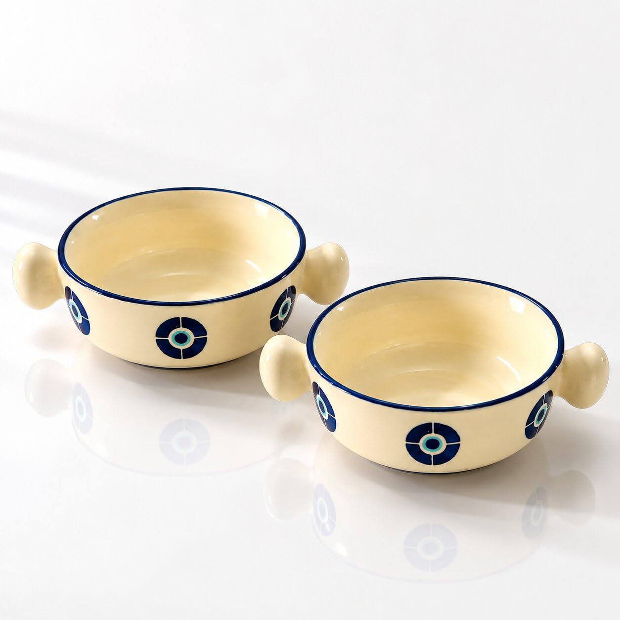 Two cream-colored ceramic bowls with side handles, decorated with blue and white circular patterns, shown on a reflective white surface.
