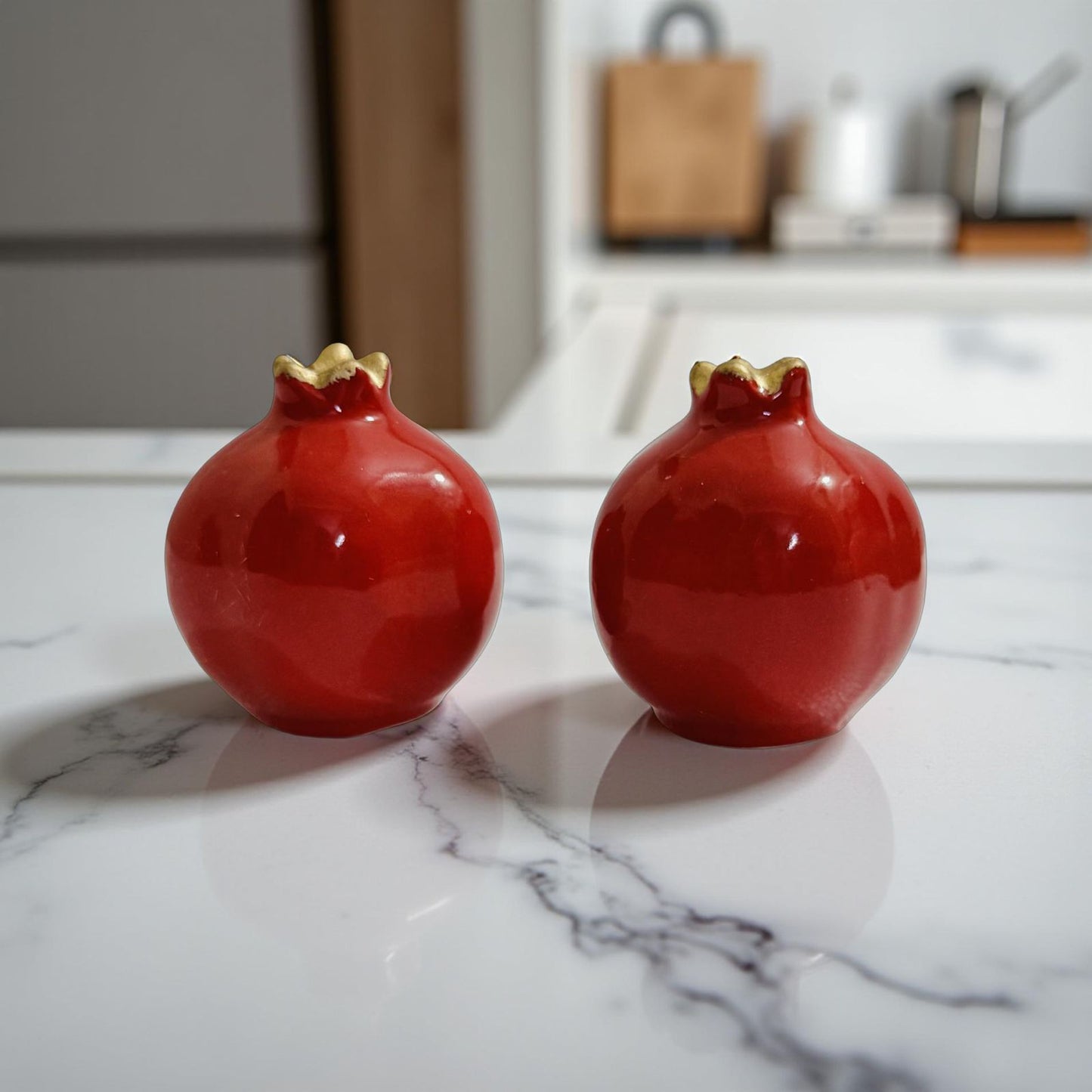 Two red ceramic pomegranate-shaped objects on a marble surface with a blurred kitchen background.