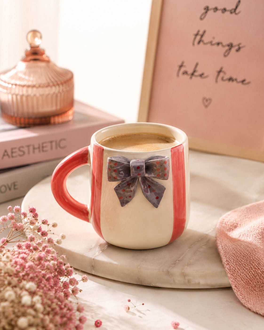 A ceramic coffee mug with a cream body, coral-red handle and side stripes, and a raised bow detail on the front, filled with coffee and styled on a marble tray with dried flowers and a pink sign in the background.