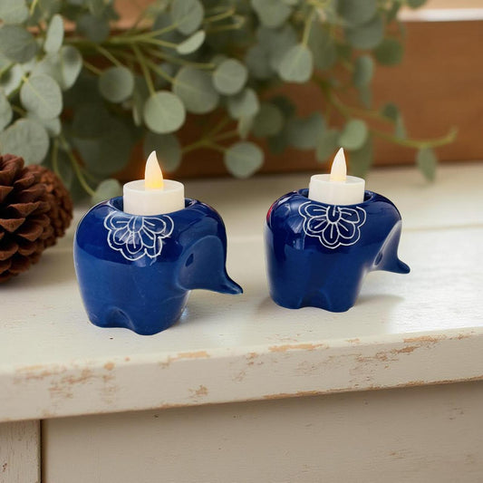 Two blue elephant-shaped candle holders with white candles on a wooden surface with greenery in the background.
