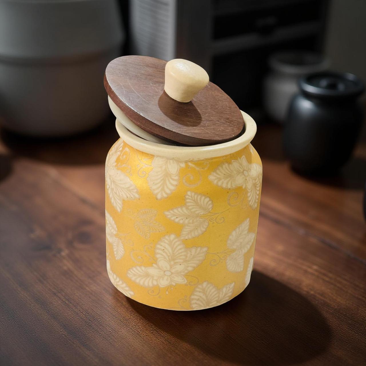 Yellow ceramic jar with leaf pattern and wooden lid on a wooden surface