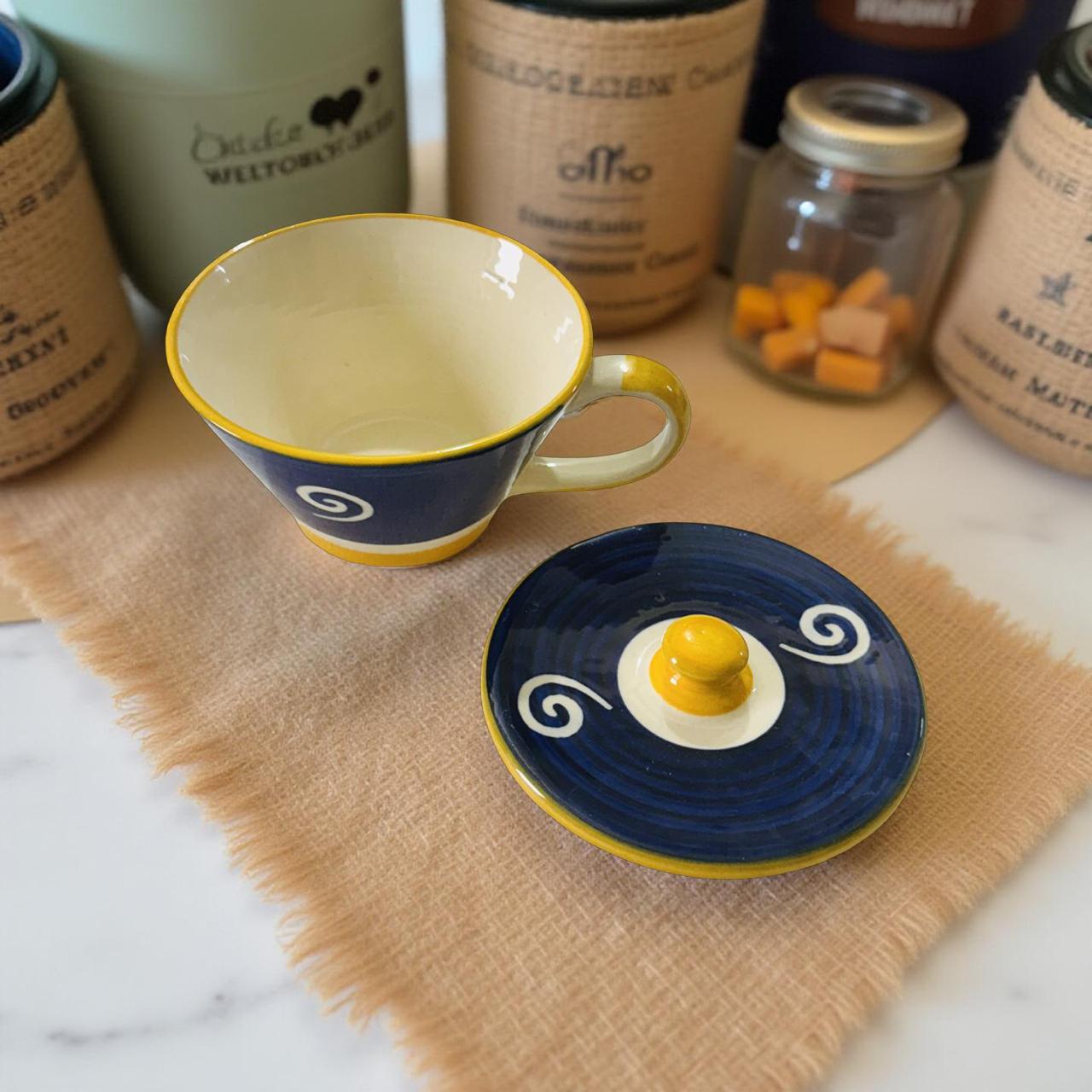 Ceramic cup with yellow interior and blue exterior, accompanied by a matching saucer with a decorative design on a textured surface.