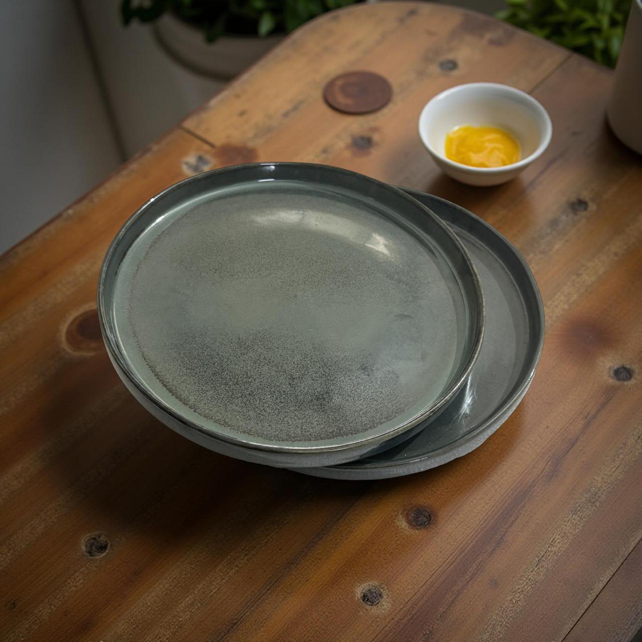 Stack of gray ceramic plates on a wooden surface with a small bowl of yellow liquid in the background.