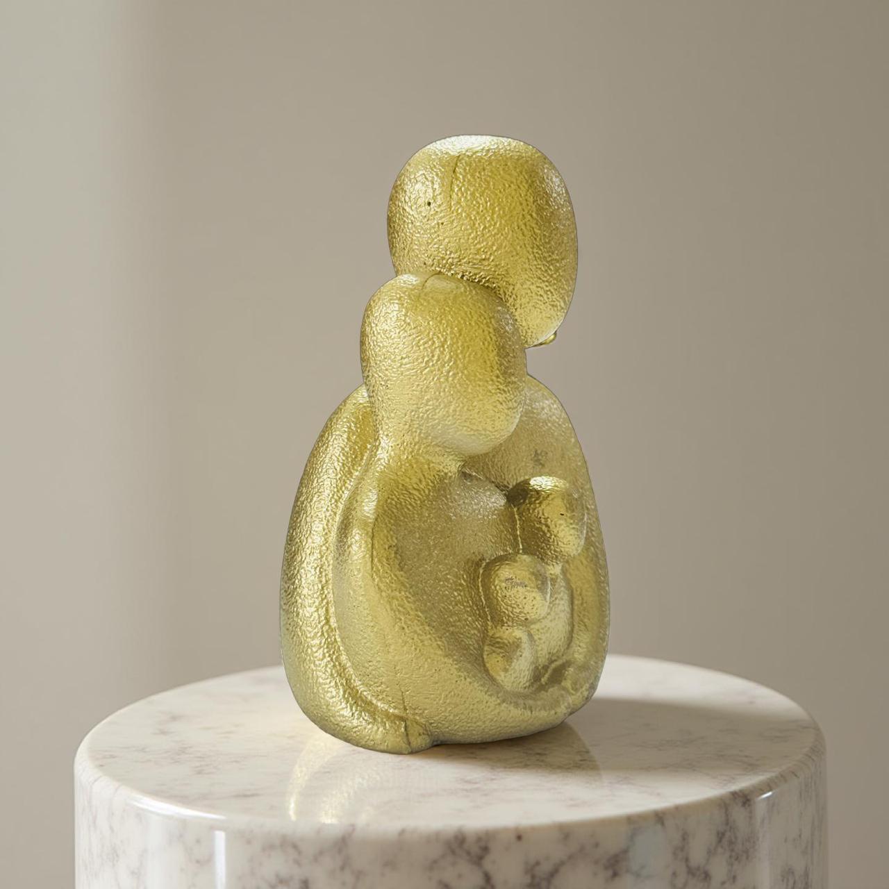 Gold sculpture of a mother holding a child on a marble pedestal against a beige background