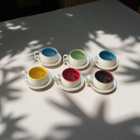 Set of six colorful ceramic cups and saucers on a light surface with shadows from leaves.