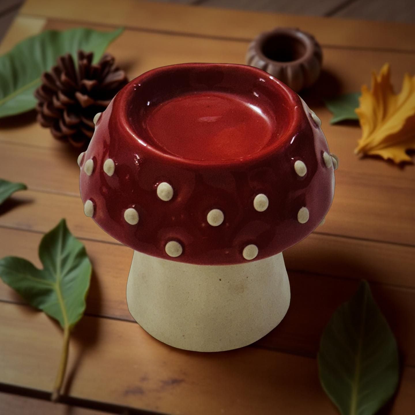 Red and white ceramic mushroom-shaped candle holder on a wooden surface with leaves and pine cones.