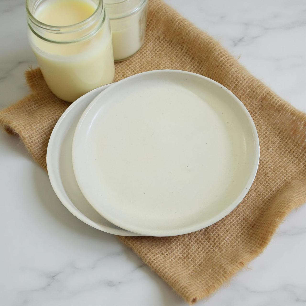 Two white ceramic plates stacked on a burlap napkin with two jars of milk in the background.