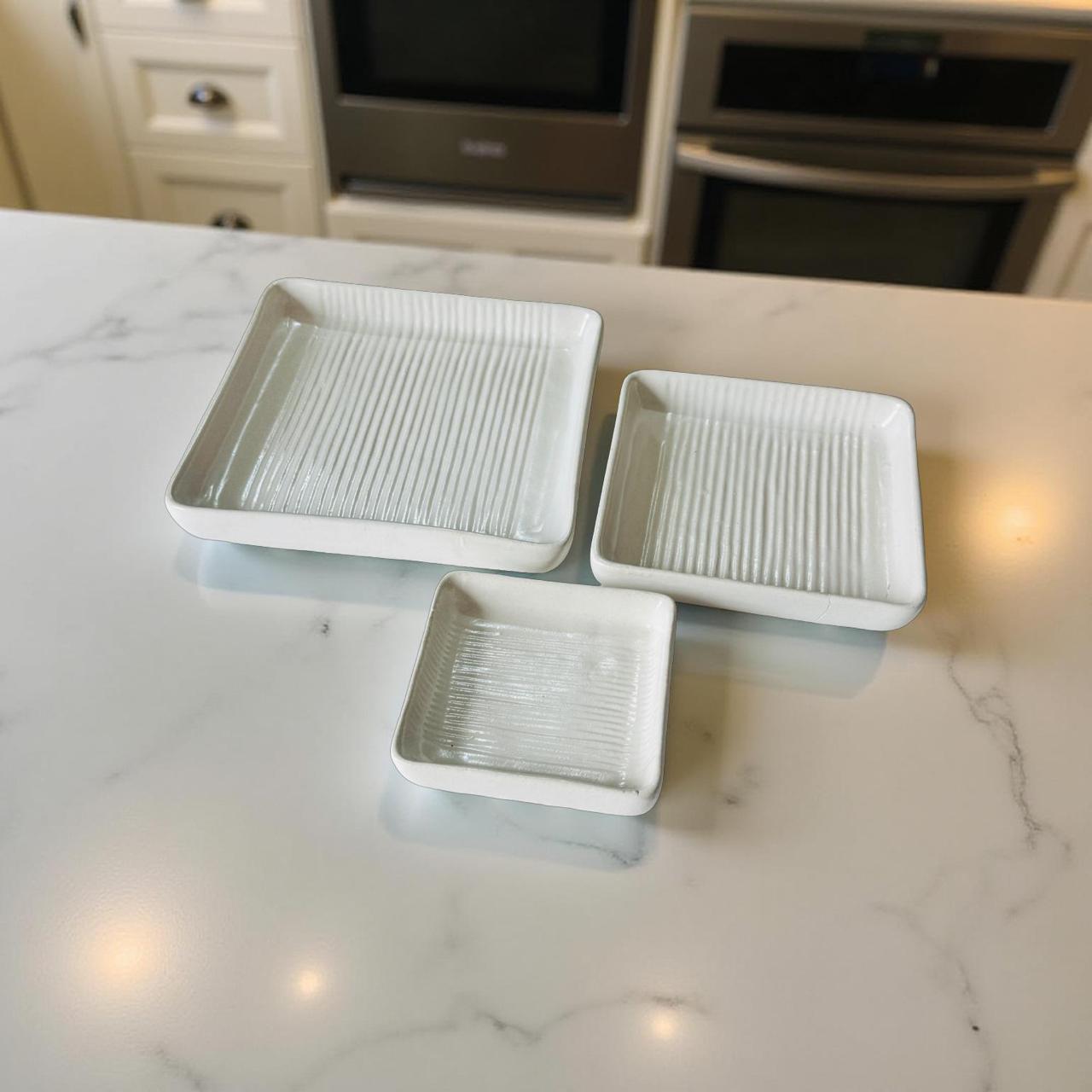 Three square ceramic trays on a marble countertop with kitchen appliances in the background.
