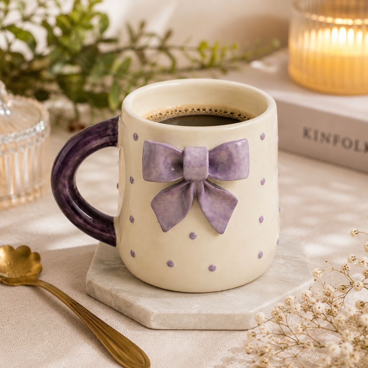 A cream-colored ceramic mug with a dark purple handle and a large purple bow decoration on the front, filled with coffee and resting on a marble coaster in a styled tabletop setting.