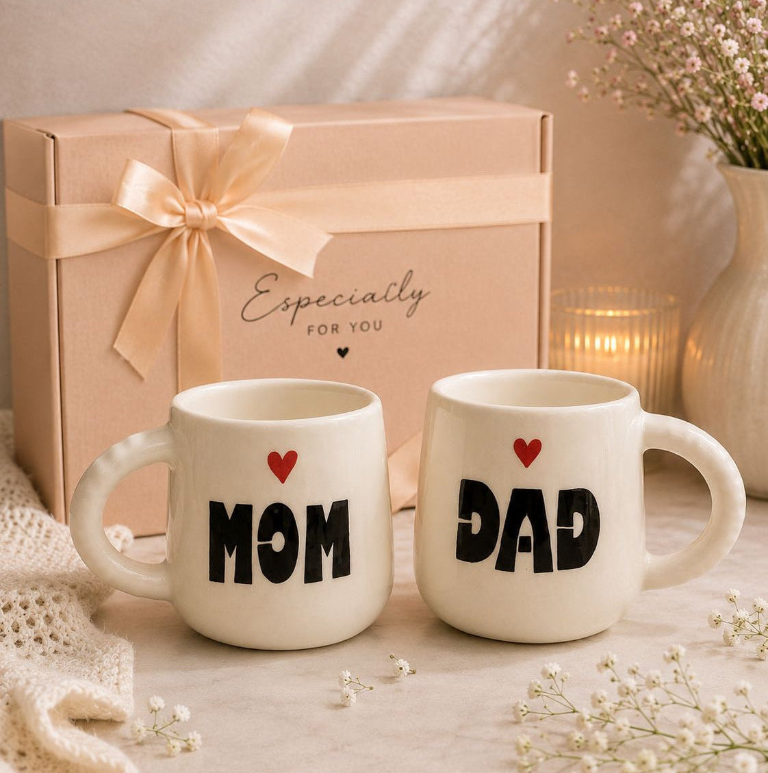 Two white ceramic mugs with black text reading 'MOM' and 'DAD' and small red hearts, displayed in front of a beige gift box with a ribbon and the words 'Especially for you.' The scene is styled with a candle, flowers, and soft warm lighting.