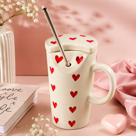 An off-white ceramic mug with a lid, a stainless steel straw, and red heart patterns, styled on a pink surface with a pink background, a book, a vase, and a small pink heart accessory.
