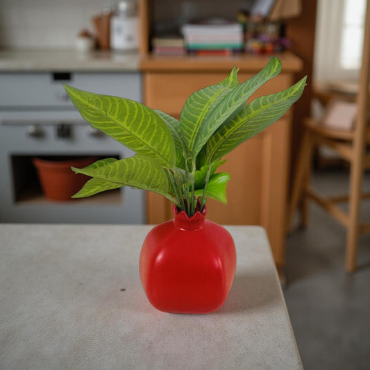 Red ceramic pomegranate vase with faux green leaves placed on tabletop.