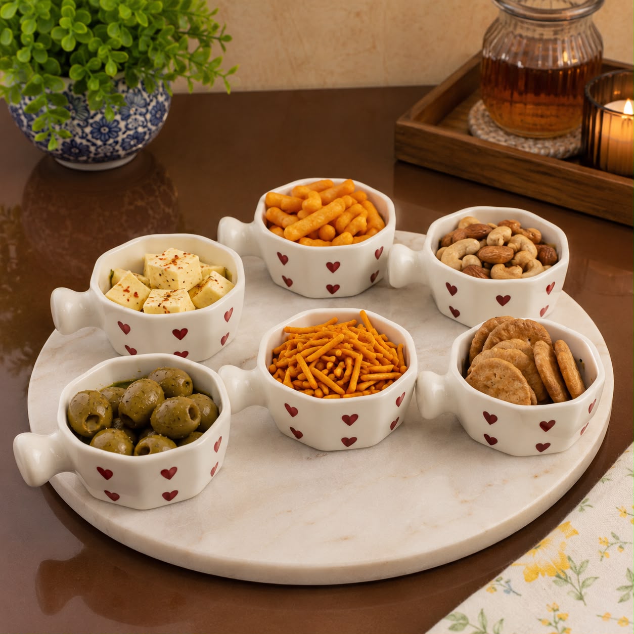 A white marble serving tray with six small white ceramic bowls decorated with red hearts, filled with snacks like crackers, cheese, olives, and nuts, set on a dark brown table with a potted plant and a candle in the background.