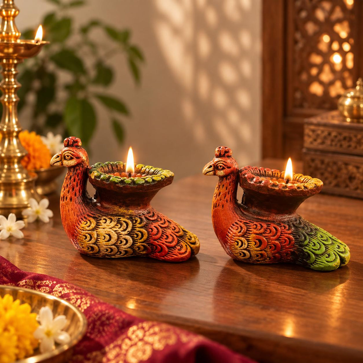 Two decorative peacock-shaped diya lamps with lit flames, placed on a wooden table in a warmly lit indoor setting with floral and brass decor in the background.