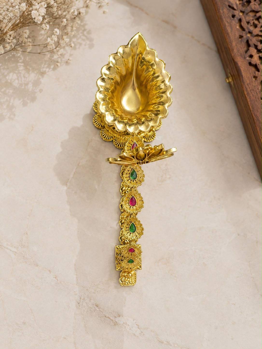 A decorative gold-toned oil lamp with a lotus-shaped top and ornate handle, accented with small red and green stones, displayed on a light marble surface.