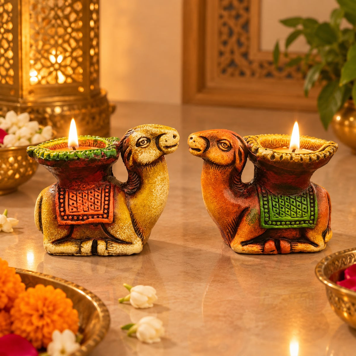 Two decorative camel-shaped diya lamps with lit candles, shown on a polished tabletop with festive flowers and warm ambient background decor.