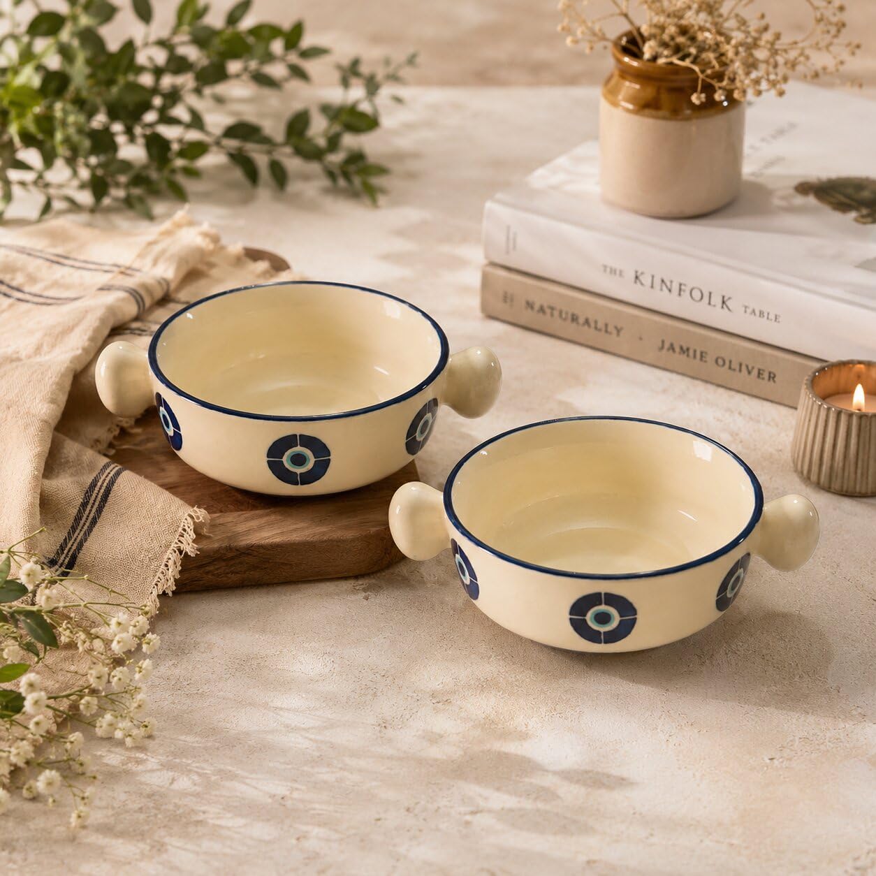 Two cream-colored ceramic bowls with blue geometric patterns and side handles, styled on a rustic beige tablecloth with books, a candle, and greenery in the background.