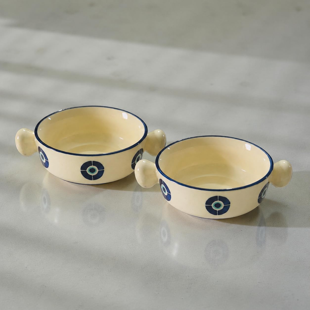 Two small cream-colored ceramic bowls with blue rims and blue circular patterns, each with a small side handle, set on a light marble surface.