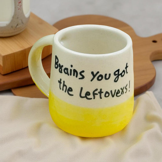 White mug with yellow bottom and text 'Brains you got the leftovers!' on a wooden surface.