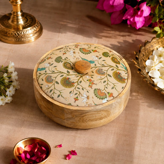 A round wooden box with a matching lid decorated with intricate floral and bird motifs in green, orange, and blue on a beige background, styled on a beige fabric surface with decorative flowers and a brass vessel nearby.
