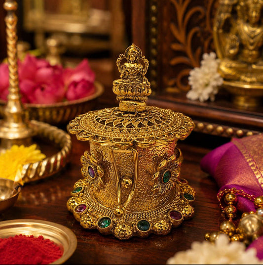 An ornate gold-toned decorative idol or lamp with a small figurine on top, set on a dark wooden surface with flowers, red powder, and other decorative items in a warm indoor setting.