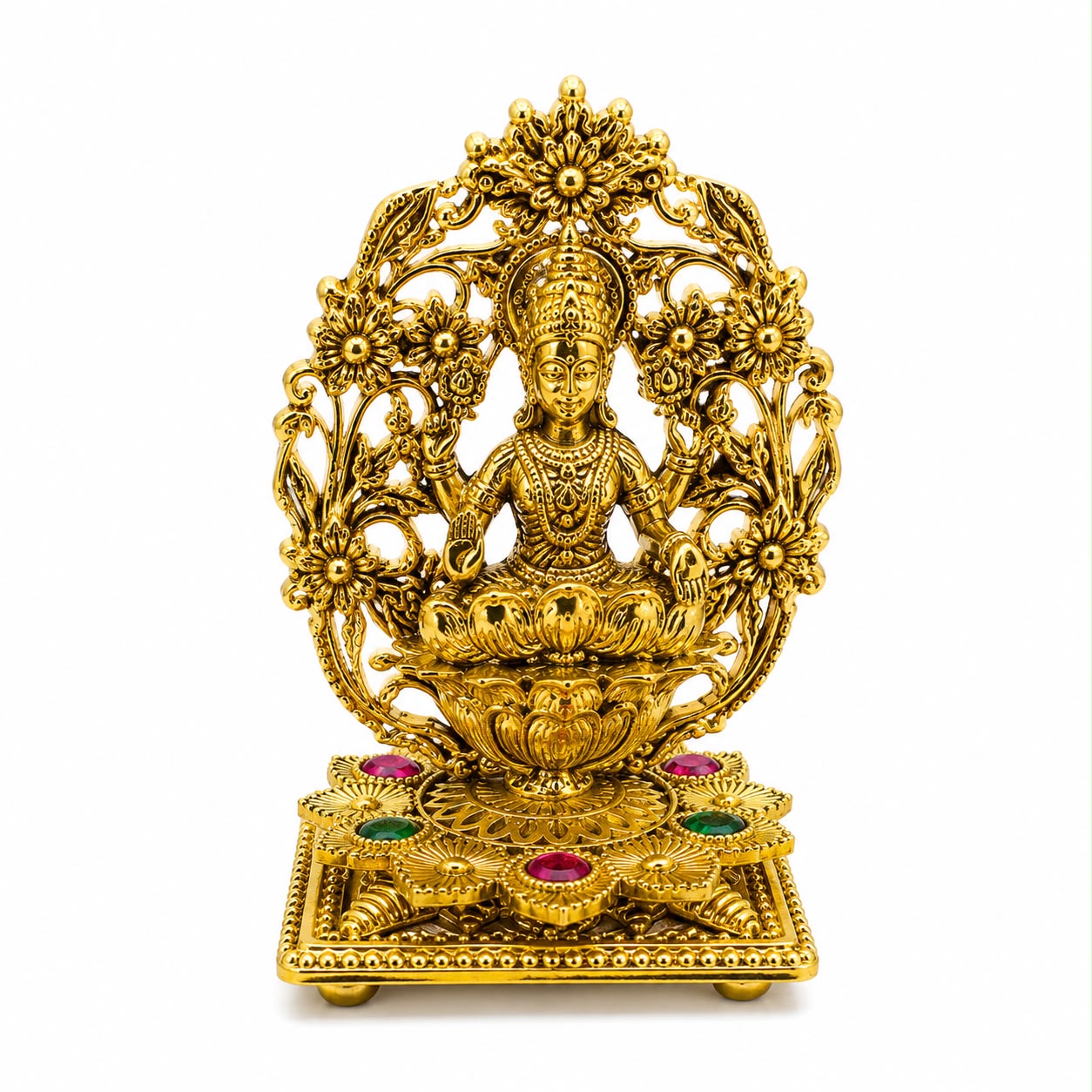 An ornate gold-toned metal statue of a seated deity on a lotus pedestal, surrounded by floral motifs and mounted on a rectangular base with small red and green jewel accents, photographed against a plain white background.