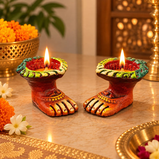 Two decorative foot-shaped diya candles with lit flames, placed on a marble-like surface with warm festive decor in the background.