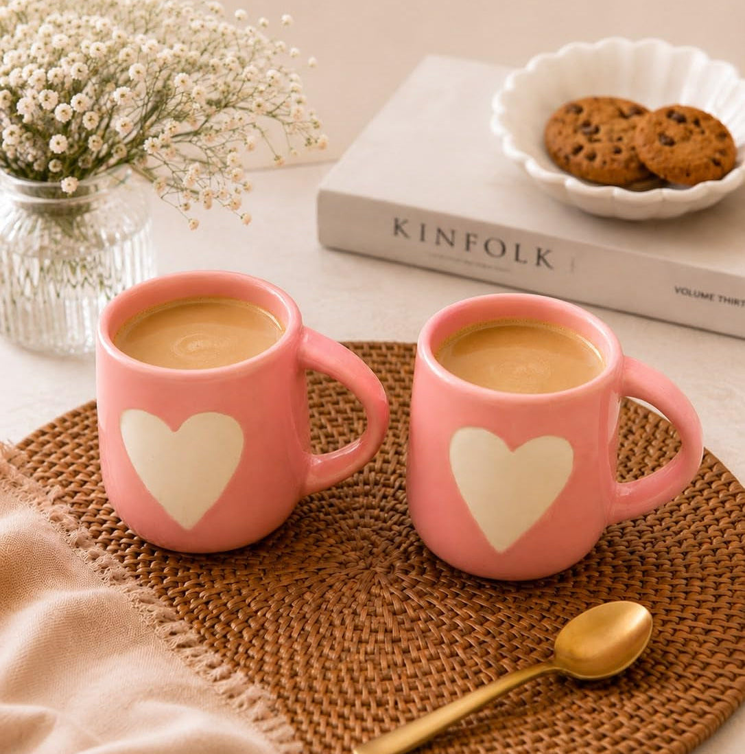 Two pink ceramic mugs with white heart designs, filled with coffee, arranged on a woven brown placemat with a gold spoon. A beige cloth, a glass vase with dried flowers, a book, and cookies are styled in the background.