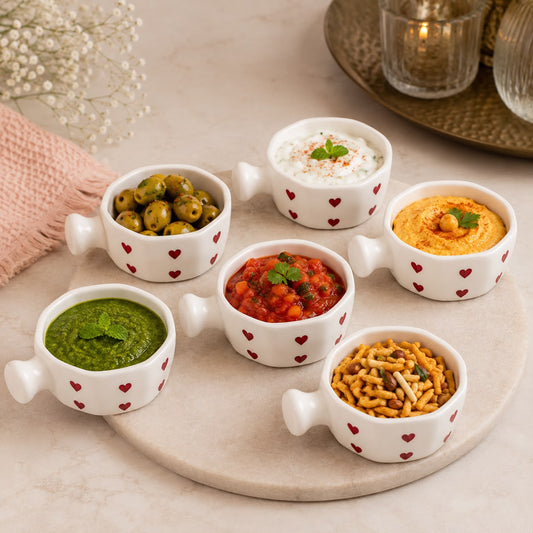 Six small white ceramic bowls with heart patterns are arranged on a round stone tray, filled with various foods like olives, dips, and snacks, set on a light marble surface.