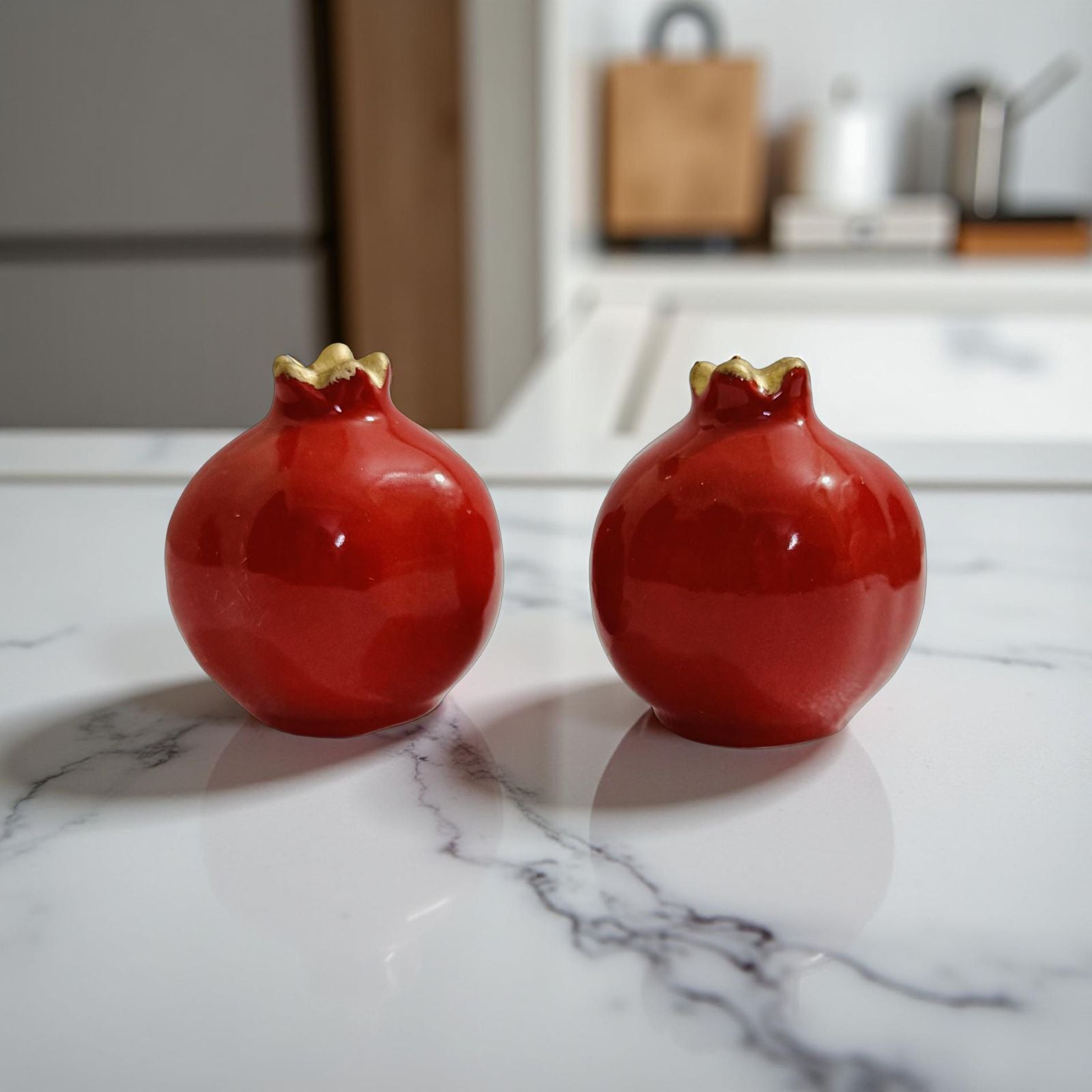 Two red ceramic pomegranate-shaped objects on a marble surface with a blurred kitchen background.