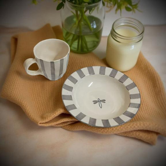 Ceramic cup and bowl with gray stripes on a beige cloth, with a glass of milk and plant in the background.