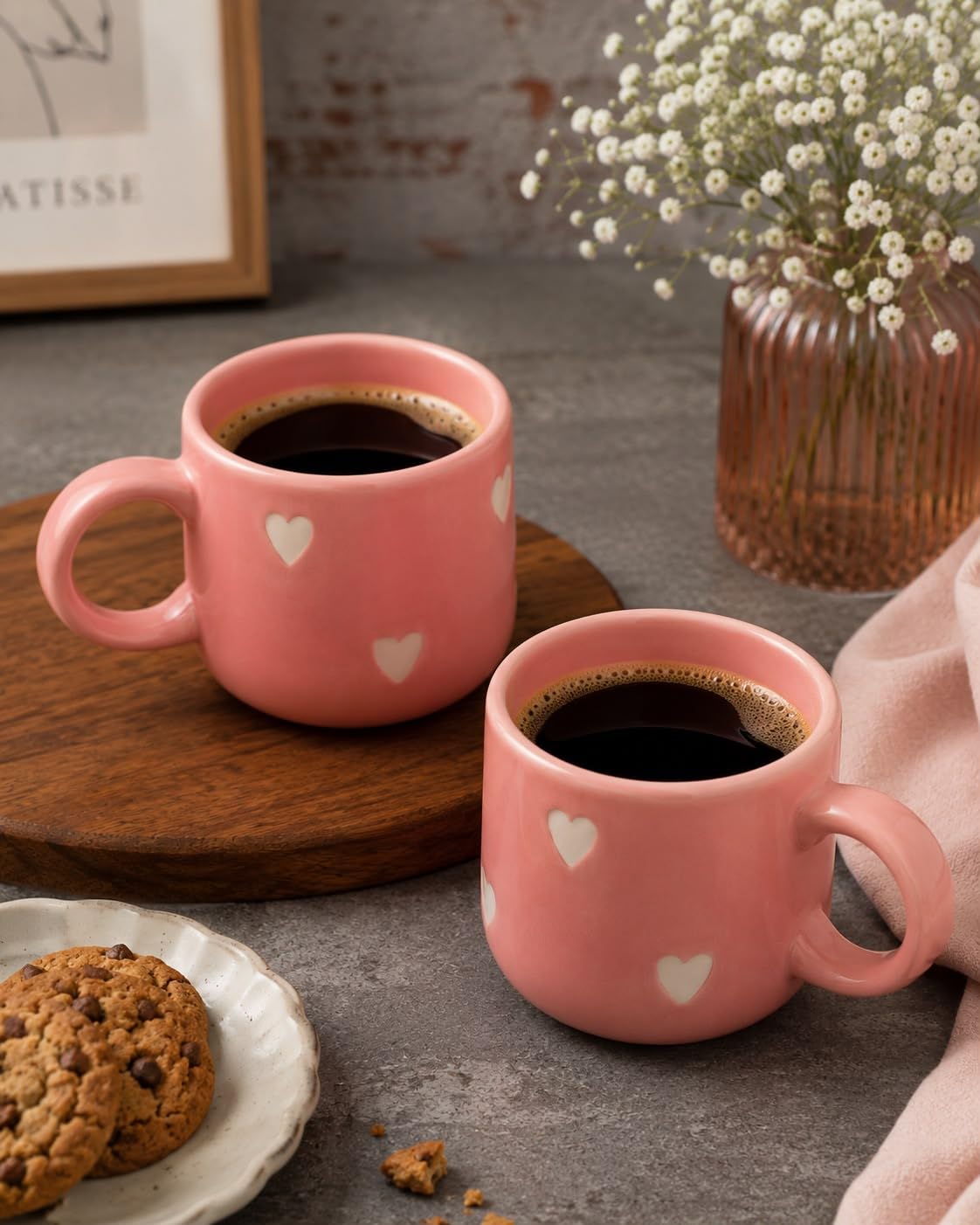 Two pink ceramic mugs filled with black coffee, decorated with small white hearts, styled on a gray tabletop with a wooden board, cookies, and a vase of white flowers in the background.