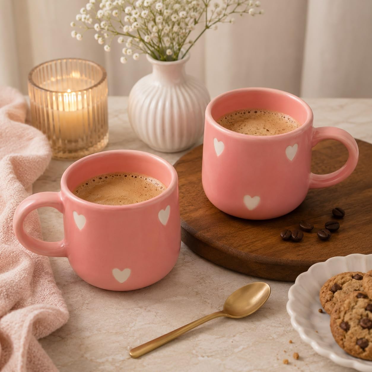 Two pink ceramic mugs with small white heart patterns, filled with coffee, styled on a table with a wooden spoon, cookies, a candle, and a vase of baby's breath.