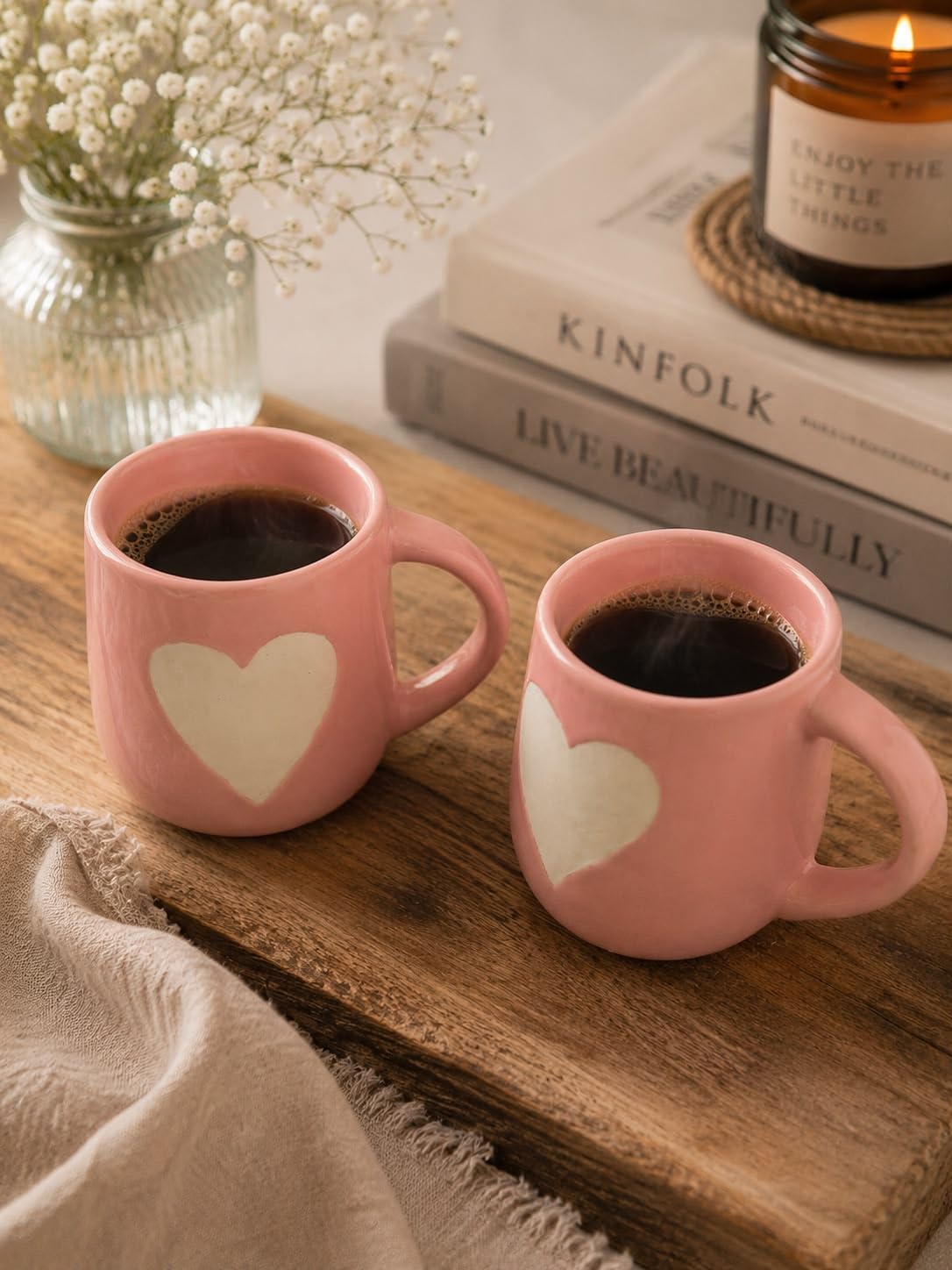 Two pink ceramic mugs with white heart designs filled with black coffee, styled on a wooden tray with books, a candle, and baby's breath flowers in the background.