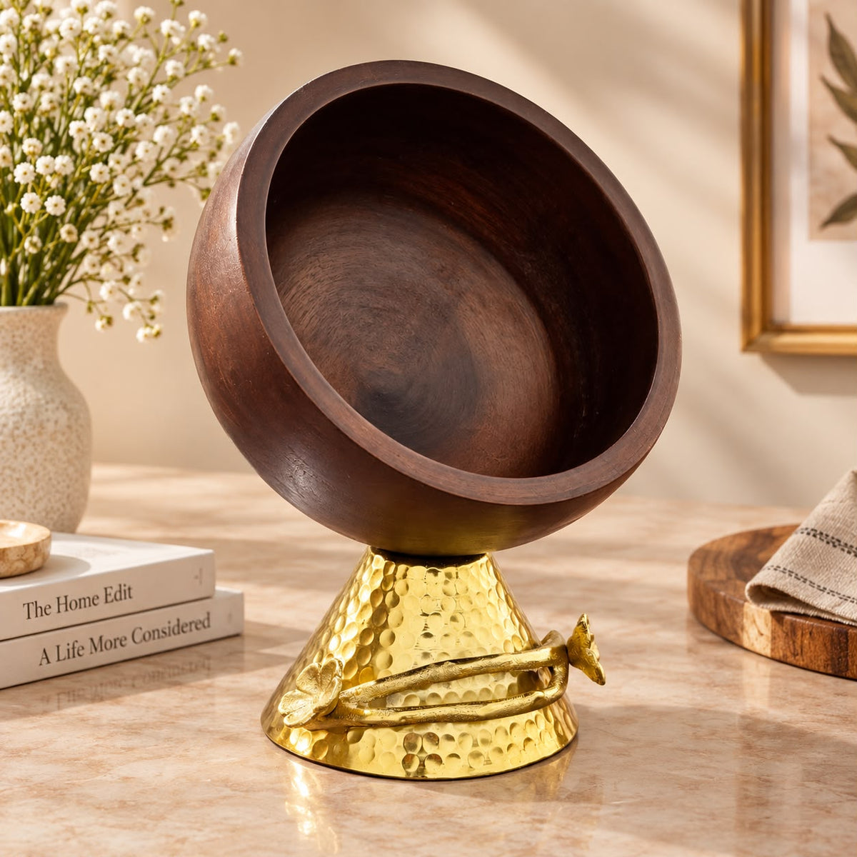 A decorative bowl with a dark brown wooden interior and a hammered gold metal base, styled on a tabletop with books, a vase of small white flowers, and a folded cloth in a warm indoor setting.