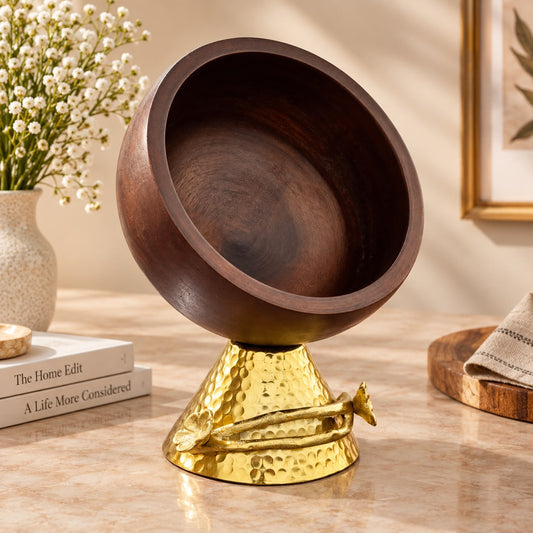 A decorative bowl with a dark brown wooden interior and a hammered gold metal base, styled on a tabletop with books, a vase of small white flowers, and a folded cloth in a warm indoor setting.