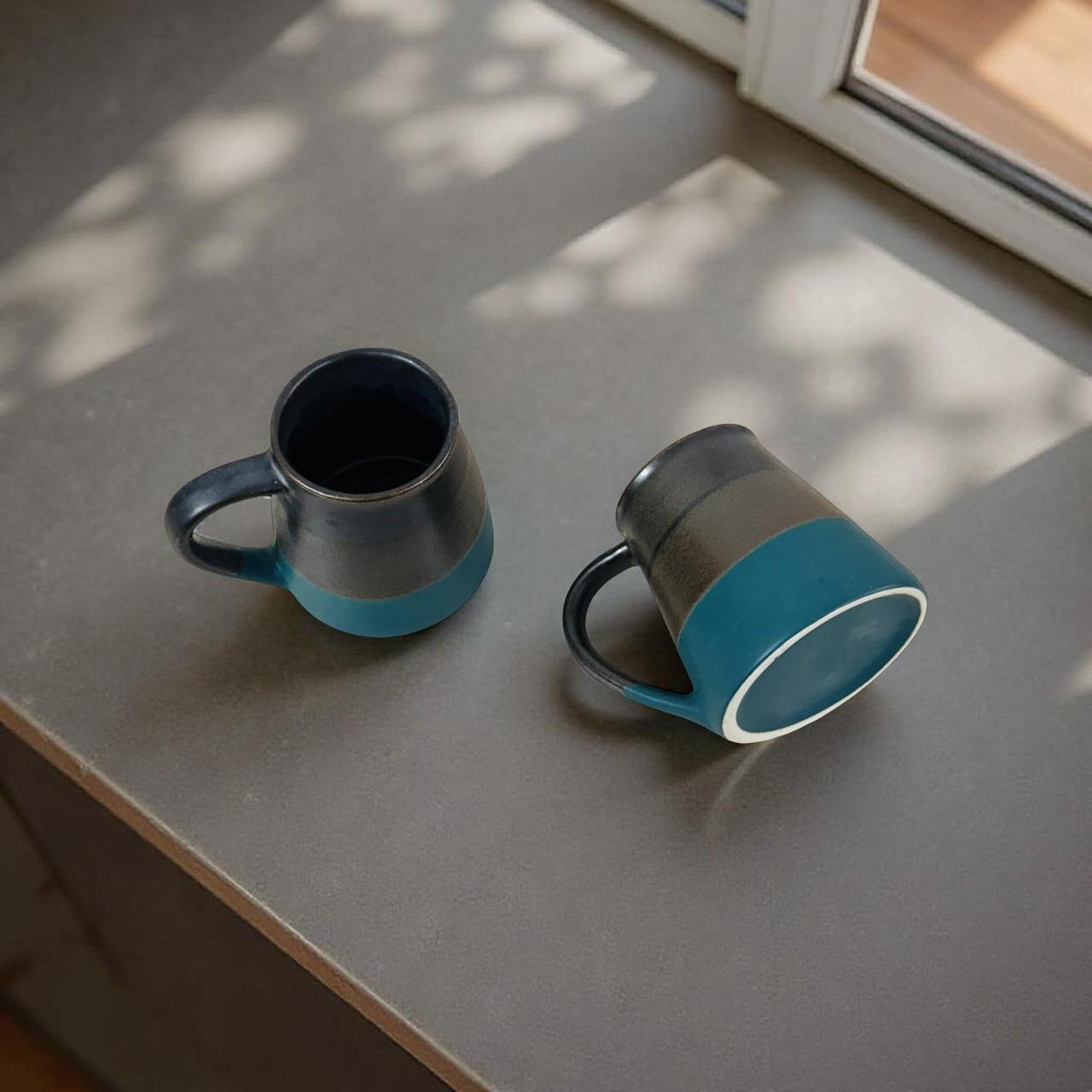 Two blue and gray ceramic mugs on a gray surface with a window in the background.