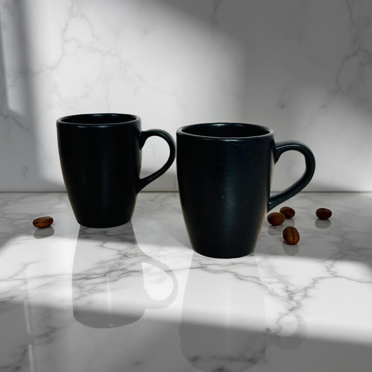 Two black mugs on a marble surface with scattered coffee beans.