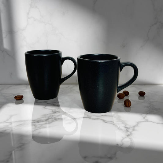 Two black mugs on a marble surface with scattered coffee beans.