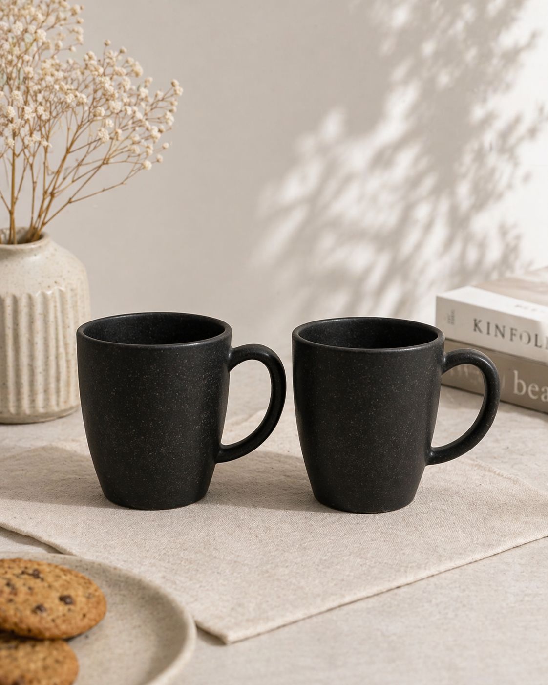 Two black mugs on a light surface with a vase, books, and cookies in the background.