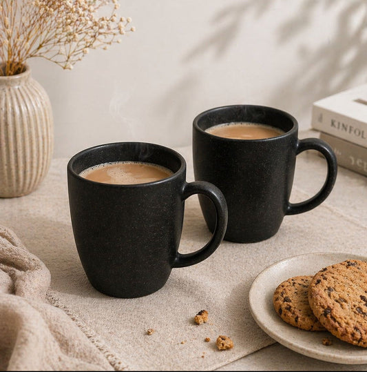 Two black mugs with coffee on a table with cookies and dried flowers.