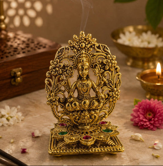 An ornate gold-toned metal incense burner figurine depicting a seated deity, with visible smoke rising from the top. The figurine is styled on a stone surface with a pink flower, a lit diya, a brass bowl, and a wooden box in a warm, atmospheric setting.