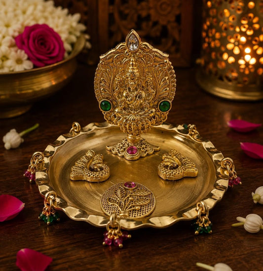 An ornate gold-toned Diwali oil lamp and matching decorative tray set on a dark wooden surface, with flowers and a glowing lantern in the background.
