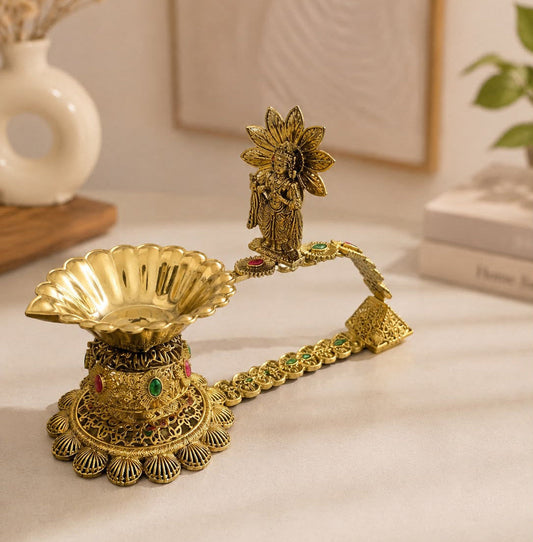 An ornate gold-toned brass oil lamp with a decorative handle, featuring a small figurine at the top and colorful stone accents, displayed on a light surface with a softly blurred home background.
