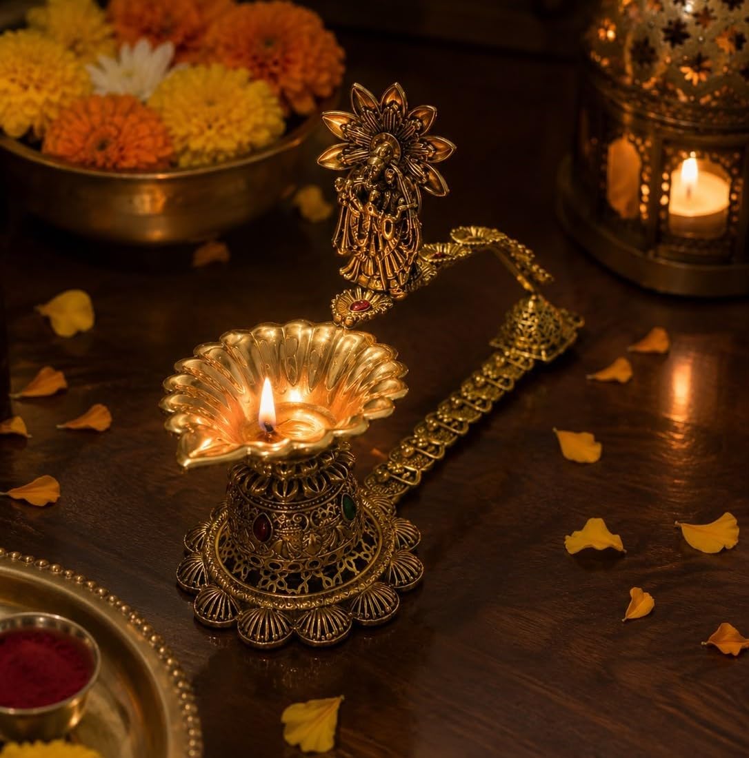 An ornate brass diya lamp with a decorative handle and a lit candle, placed on a dark wooden table with scattered flower petals, a bowl of marigolds, and another lantern in the background.