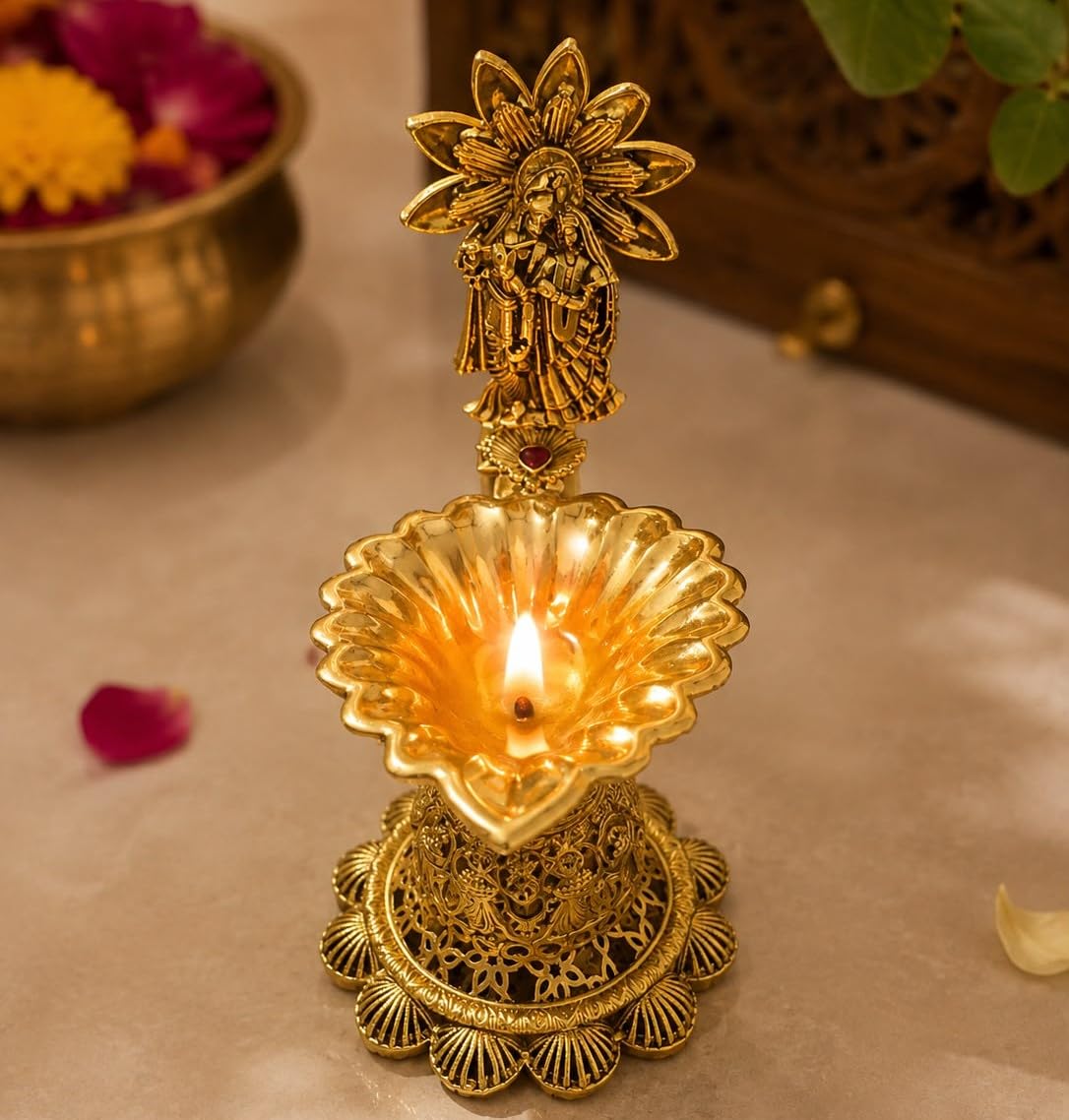 A brass oil lamp (diya) with a decorative figurine on top, lit with a warm flame, set against a softly blurred background with floral elements.