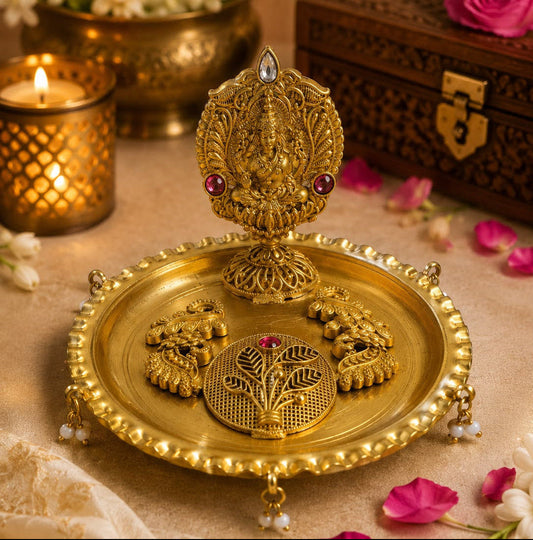 An ornate gold-toned decorative brass plate with a raised idol, shown on a draped beige fabric with scattered pink and white flowers, a lit candle, and a carved wooden box in the background.