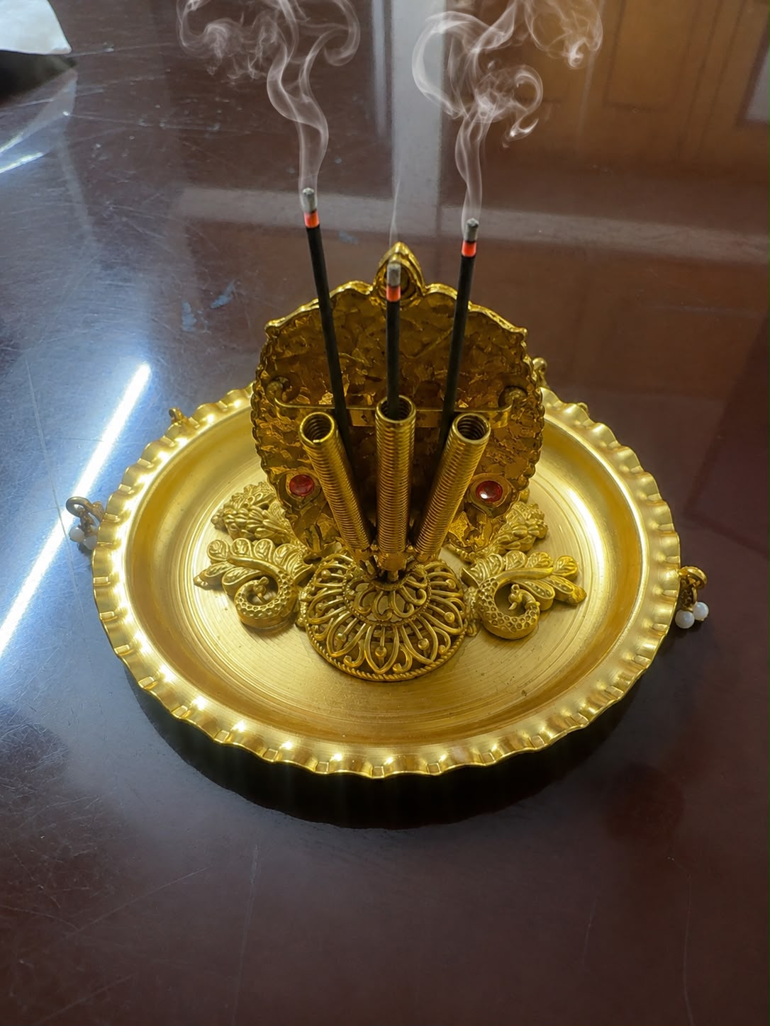 A decorative gold incense holder with three incense sticks standing upright, smoking, placed on a reflective dark brown surface.