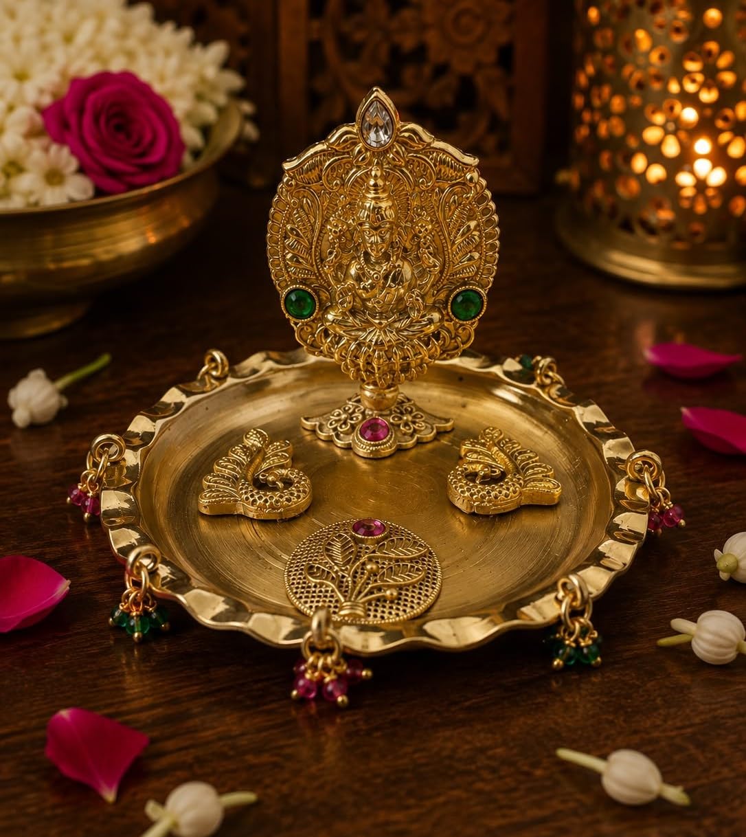 An ornate gold-toned brass diya lamp set on a dark wooden surface, featuring a central deity lamp, three peacock-shaped lamps, and a round decorative plate with hanging red and green jewel accents. The background includes a warm glowing brass lantern and blurred flowers.