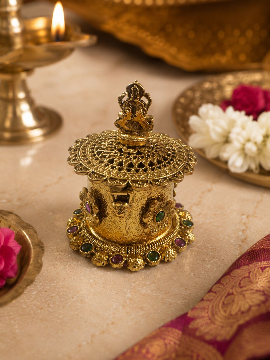 An ornate gold-toned brass idol of Lord Ganesha on a decorated base, styled on a marble surface with a lit diya, flowers, and a silk cloth in the background.